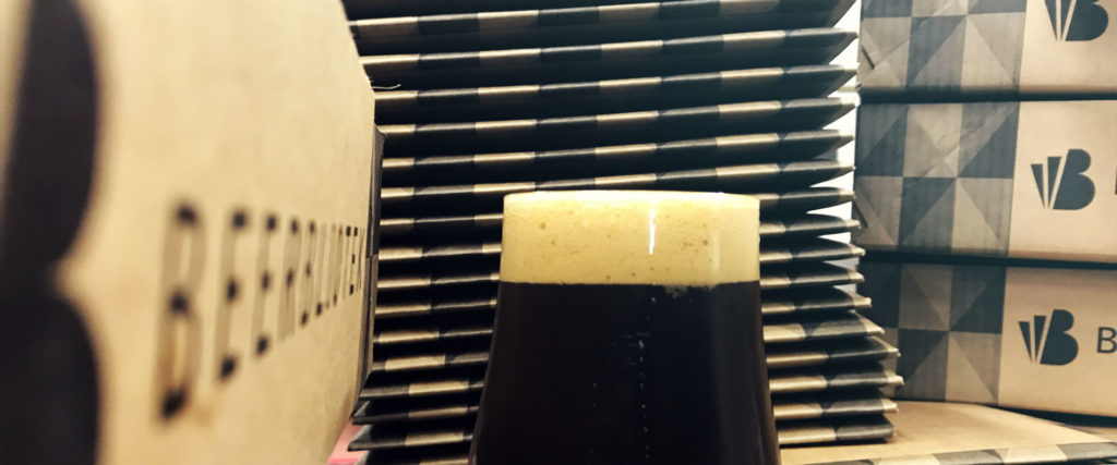 A beer glass of freshly poured, freshly packed, Beerbliotek Cascadian Dark Ale, called "No Style without Substance". On the table with packaging boxes for cans.